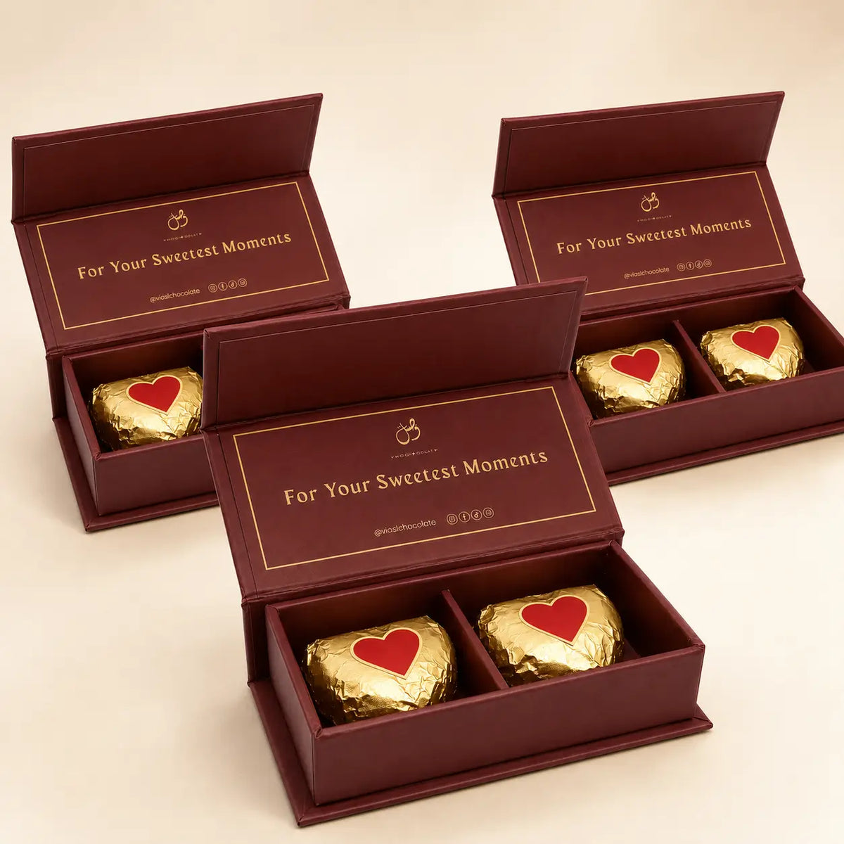 Three open dark burgundy gift boxes arranged on a light beige background, each containing two heart-shaped chocolates wrapped in shiny gold foil with a red heart on top. The inside lid of the boxes reads 'For Your Sweetest Moments' with a small crown logo.