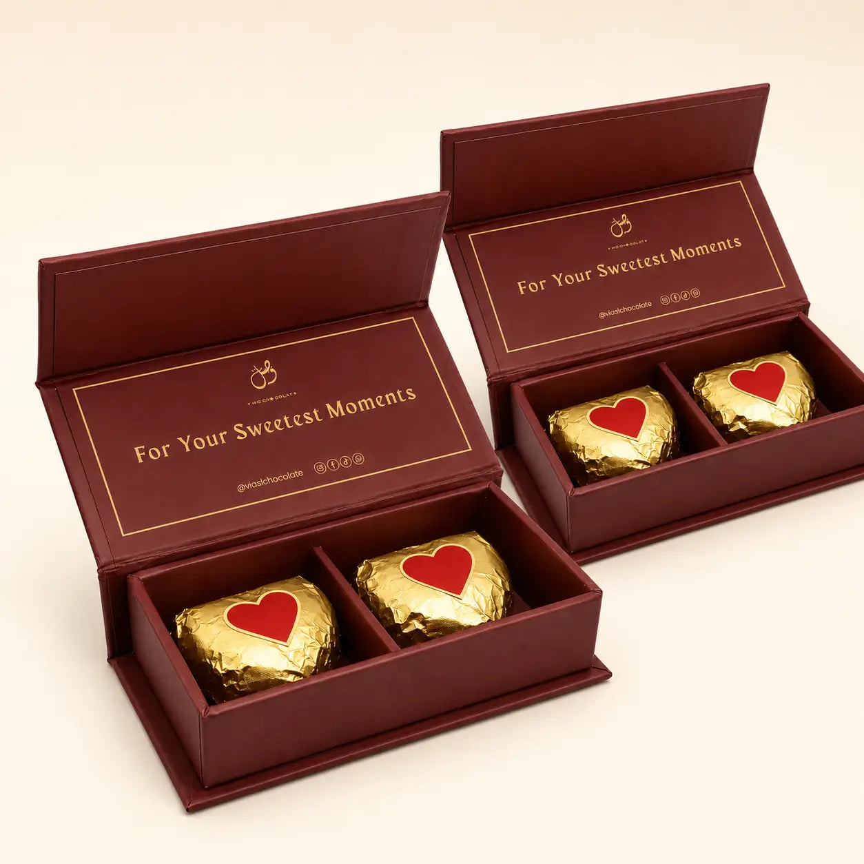 Two open burgundy gift boxes containing three gold-wrapped chocolates with red heart details, set against a light beige background.