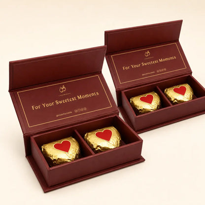Two open burgundy gift boxes containing three gold-wrapped chocolates with red heart details, set against a light beige background.